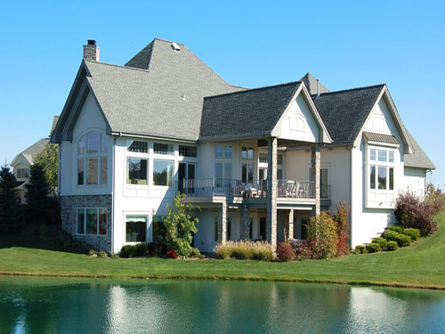 Luxury lakeside house with large windows and lush garden reflecting on water under a clear blue sky