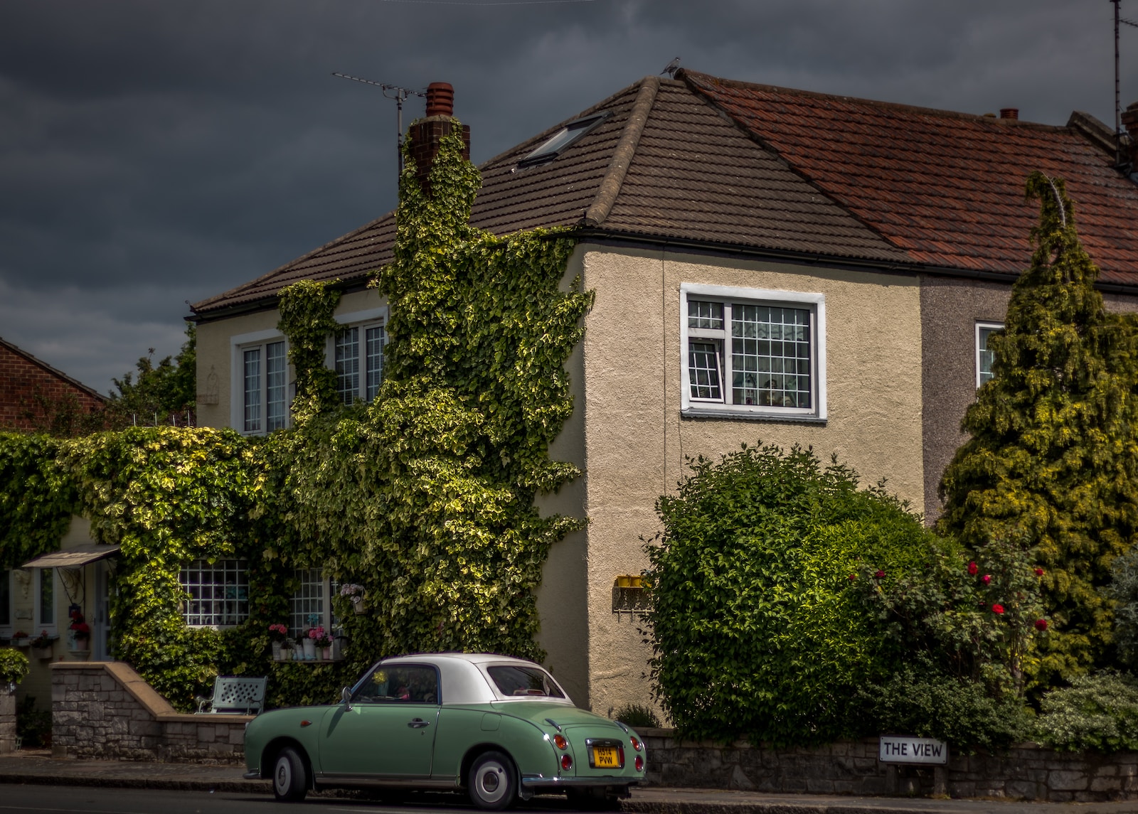 Quaint house covered in ivy with vintage green car parked outside under a cloudy sky