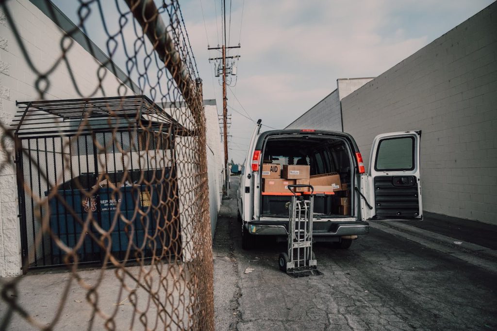 Delivery van with open doors loaded with boxes in alleyway, next to a fenced enclosure and utility pole