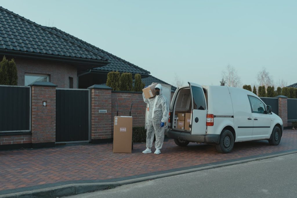 A courier in protective gear delivers packages from a van to a residential house