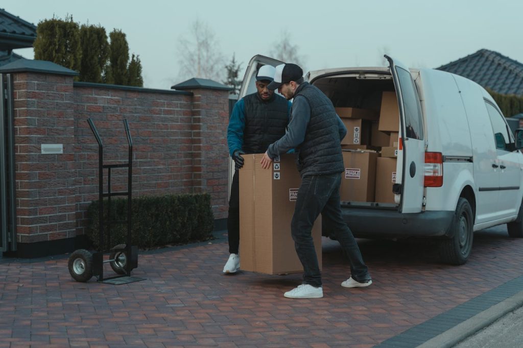 Two movers with a dolly unload cardboard boxes from a van outside a brick wall building