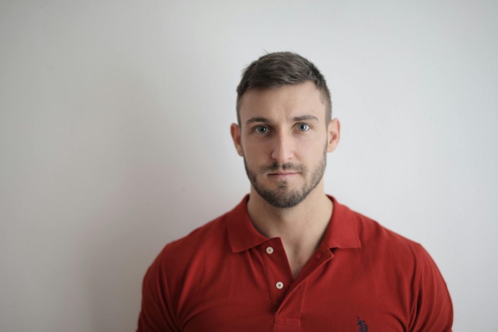A serious man in a red polo shirt poses against a white wall.